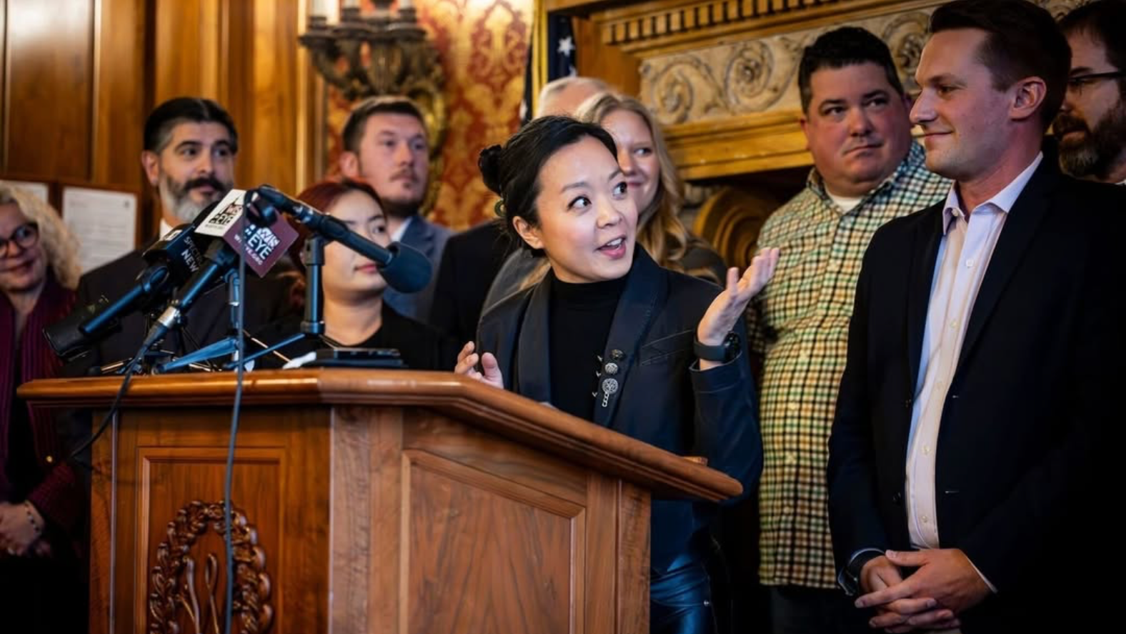 Francesca Hong speaks at podium, smiling and gesturing with one hand while addressing group of nine people standing behind her in ornate room with wood paneling