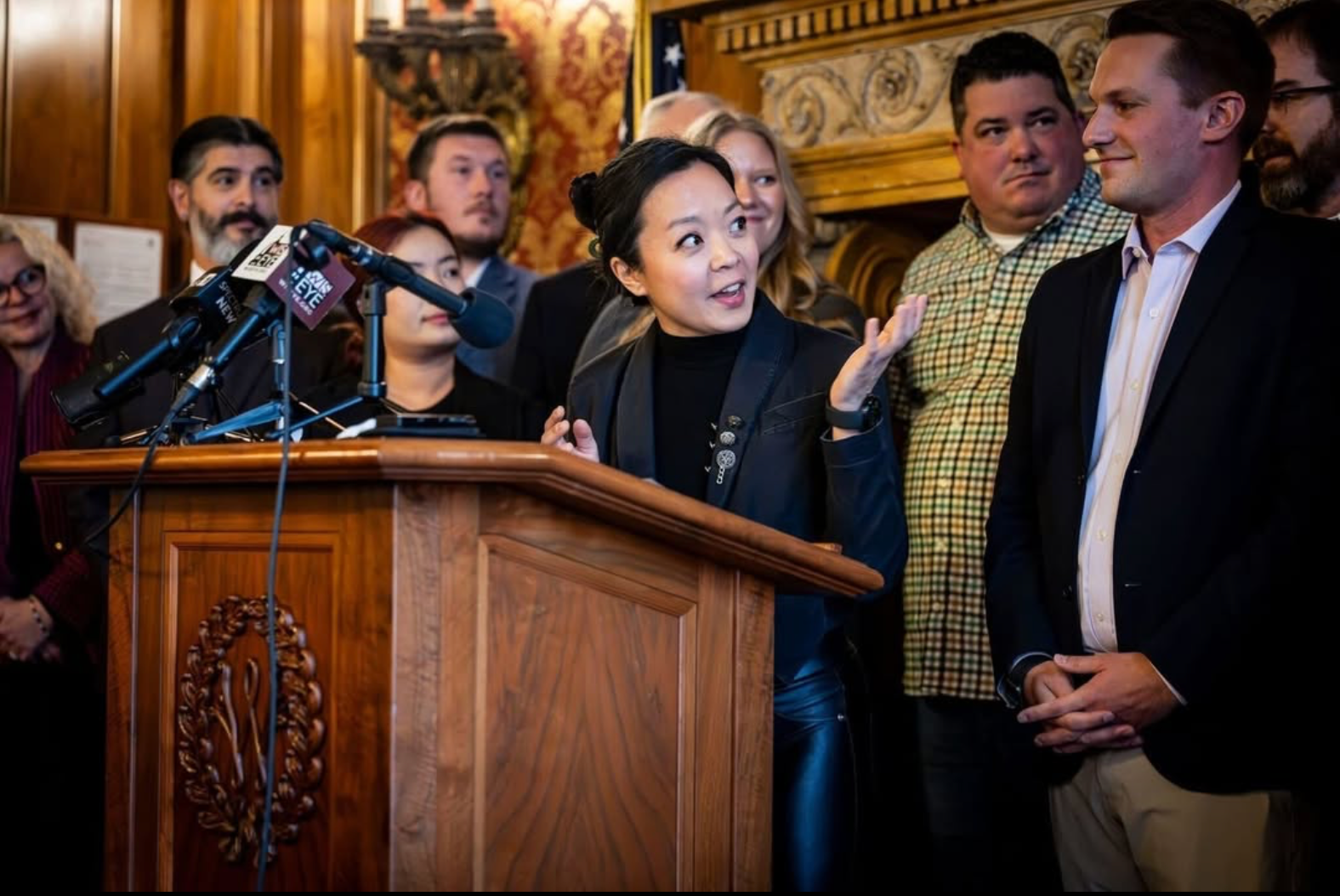 Francesca Hong speaks at podium, smiling and gesturing with one hand while addressing group of nine people standing behind her in ornate room with wood paneling